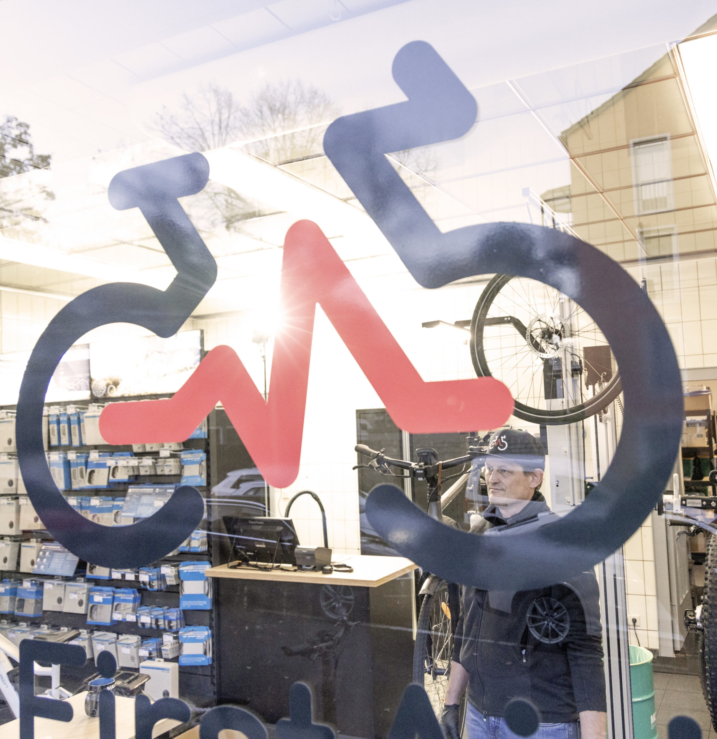 Fahrradladen-Schaufenster mit großem, stilisiertem Fahrradlogo, durch das die Werkstatt und ein Mitarbeiter im Hintergrund zu sehen sind.